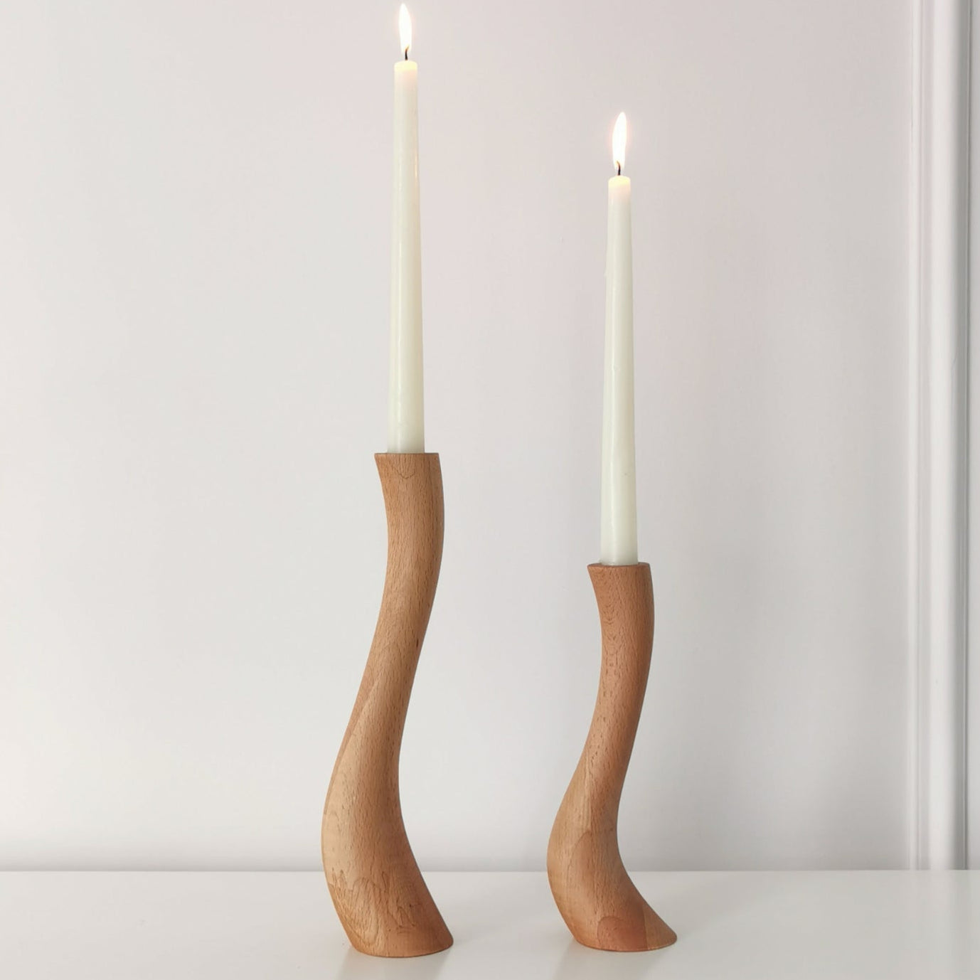 Two wooden candle holders with a Cobra-like curved design, holding white candles, placed on a white surface.